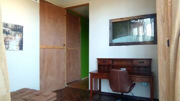 Una habitación con un escritorio de madera, una silla, un ventanal pequeño y una lámpara fijada a la pared.