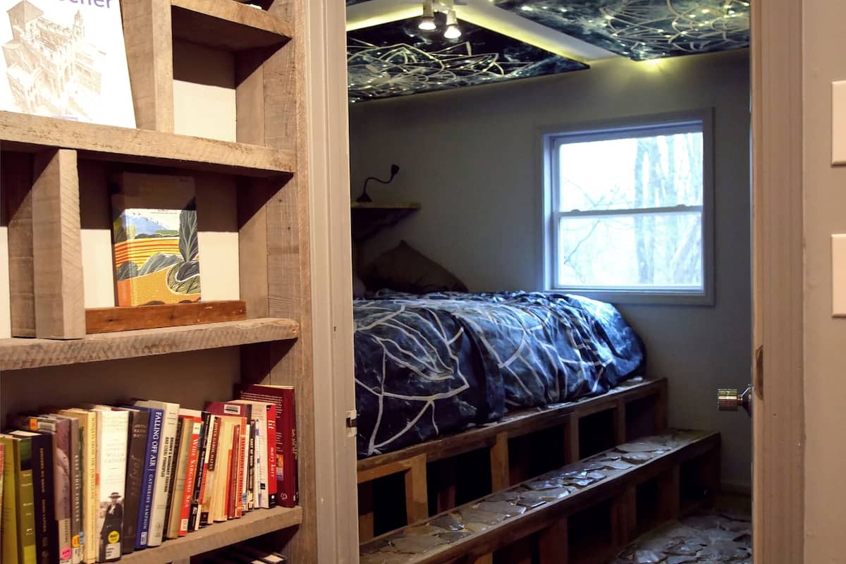 A cozy reading nook with a stone floor, a bookshelf filled with books, and a bed with a blue blanket.