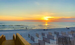 A beach with a wooden boardwalk leading to the water, a sunset in the background, and a few people relaxing on the sand.