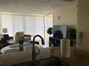 A modern living room with a sofa, a TV, a dining area with a table, and a kitchen with a sink and countertop.