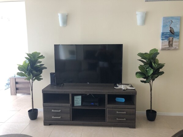Una sala de estar con una televisión de pantalla plana sobre un mueble de madera, dos plantas en macetas, un cuadro en la pared y un estante pequeño con un objeto decorativo.