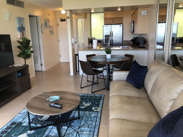 A living room with a sofa, a coffee table, and a TV stand. There is a dining area with a table and chairs. The kitchen is visible in the background with a refrigerator and cabinets.