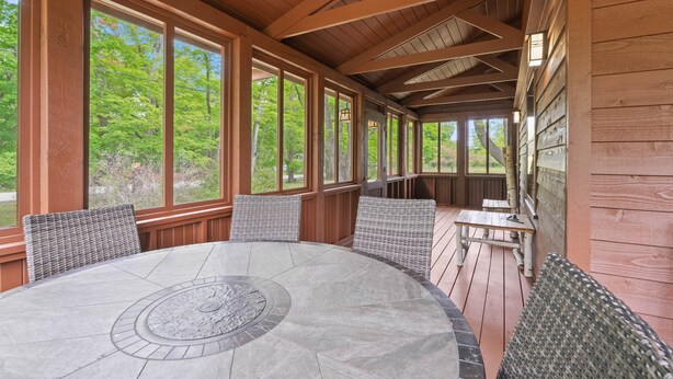 A covered outdoor seating area with a round table and chairs, large windows, and wooden beams.