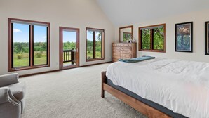 A bedroom with a large bed, a dresser, a mirror, and multiple windows offering a view of greenery.