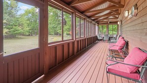 A covered porch with wooden flooring and railings, featuring a row of metal chairs with cushions, a clock on the wall, and a view of a wooded area.