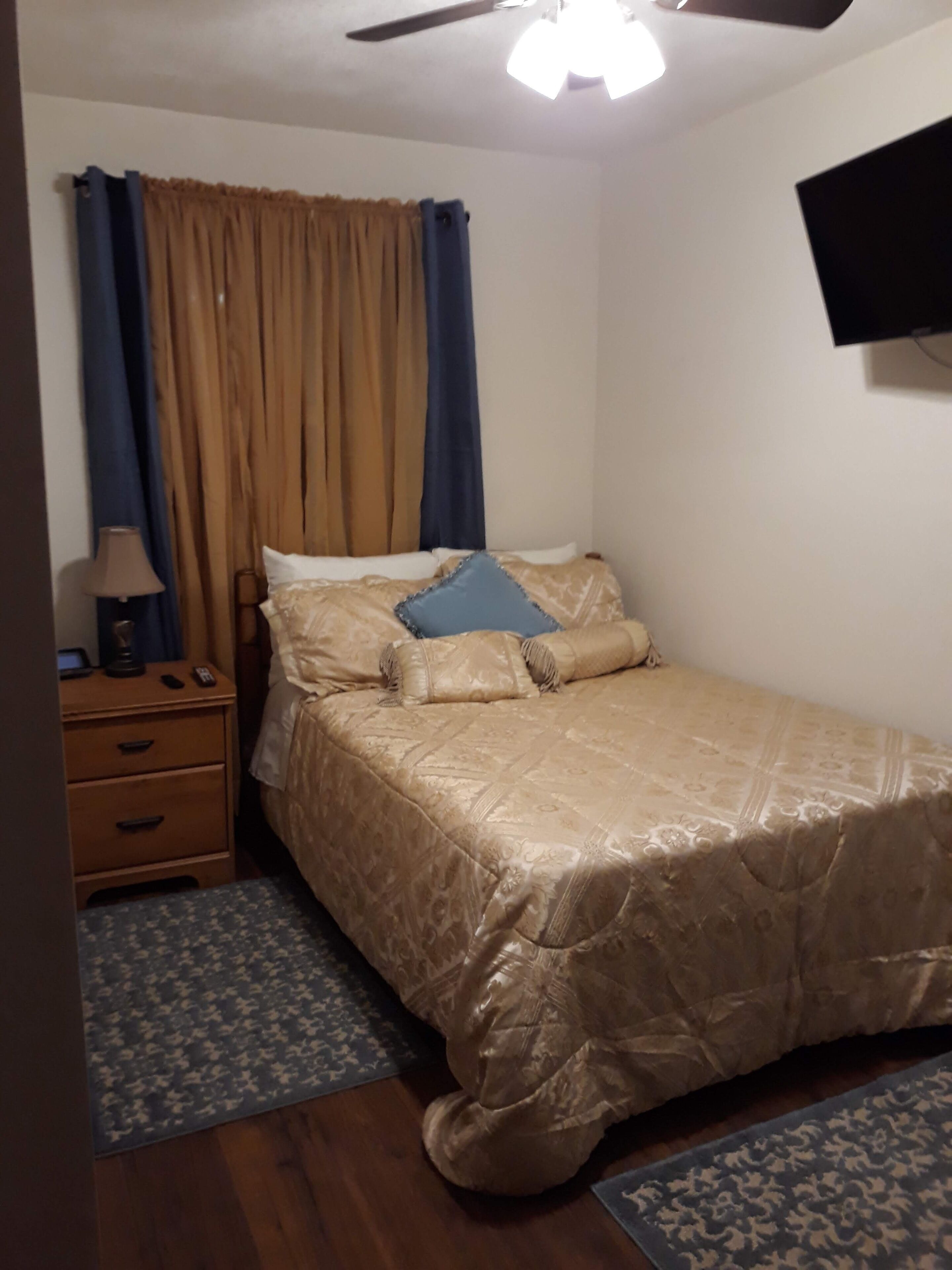 A bedroom with a bed, a nightstand, a lamp, a ceiling fan, and a TV mounted on the wall.