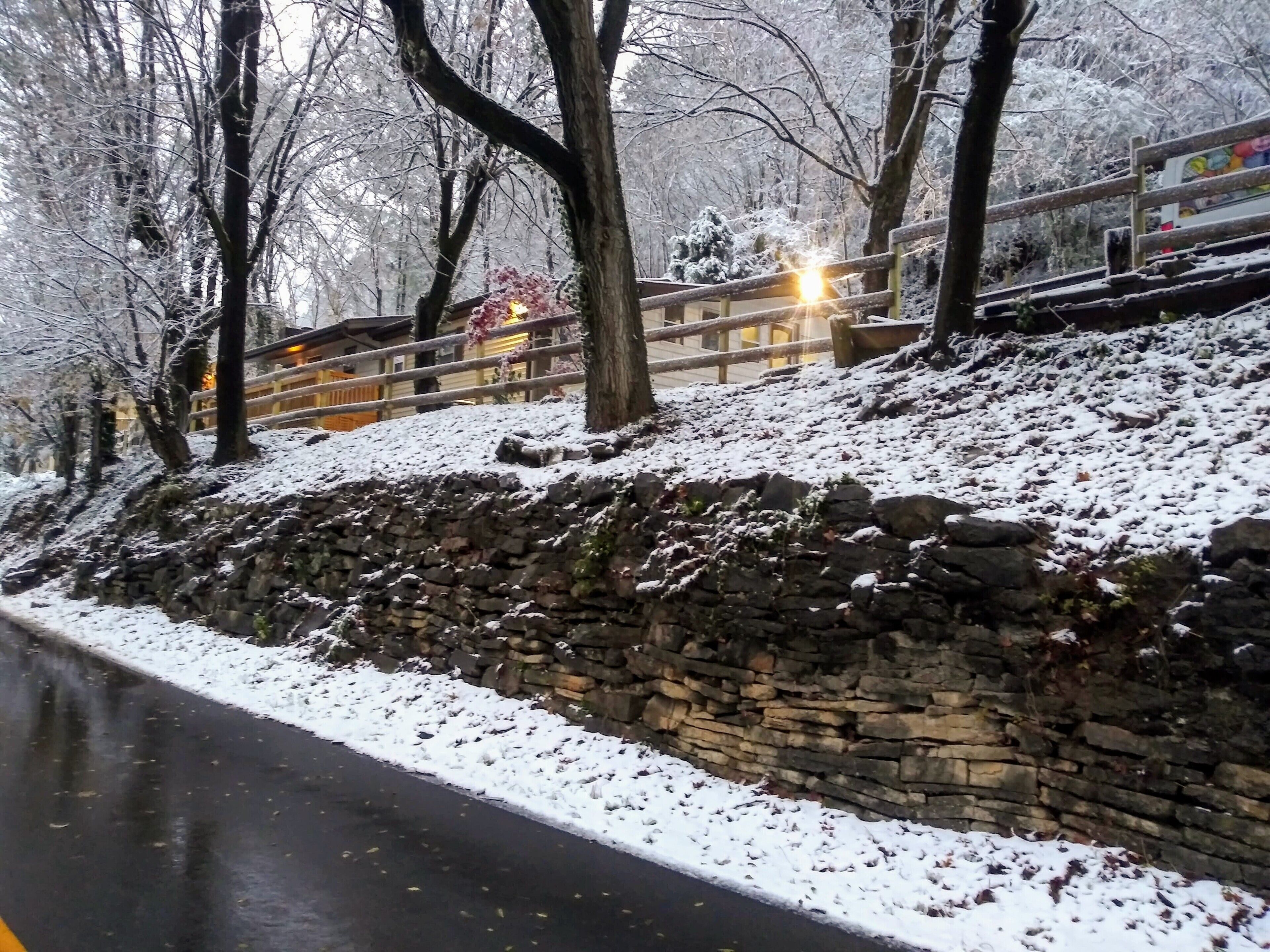 Un mur de pierre au bord d’une route, avec de la neige au sol et des arbres.