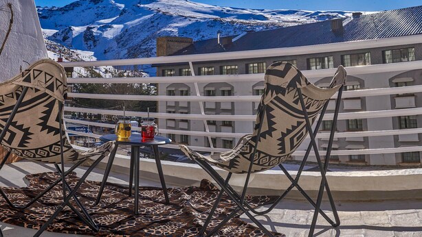Ein Balkon mit zwei gemusterten Stühlen, einem kleinen Tisch mit Getränken und Blick auf schneebedeckte Berge sowie ein Gebäude im Hintergrund.