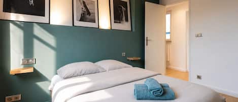 A bedroom with a bed, a nightstand, a wall-mounted light, and a door leading to another room.