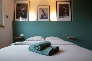 Un lit bien fait, avec du linge de lit blanc et deux serviettes enroulées, un mur d’accent de couleur turquoise et trois photographies encadrées au-dessus du lit.