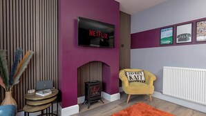 A living room with a TV mounted on a purple wall, a yellow armchair, a fireplace, and a small table with a laptop.