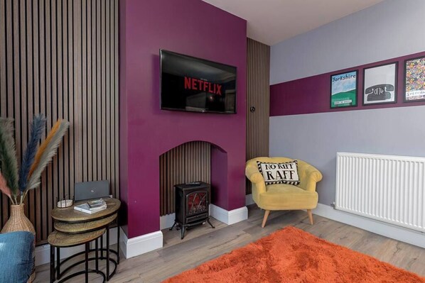 A living room with a TV mounted on a purple wall, a yellow armchair, a fireplace, and a small table with a laptop.