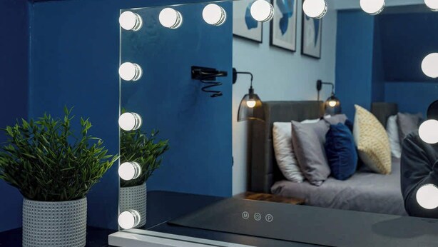 A bathroom with a mirror surrounded by lights, a potted plant, and a bed with pillows in the background.