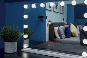 Un baño con un espejo rodeado de luces, una planta en maceta y una cama con almohadas al fondo.
