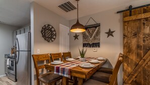 Un comedor con una mesa de madera dispuesta para cuatro personas, rodeada de sillas. Una araña cuelga encima, y se observan elementos decorativos en la pared.