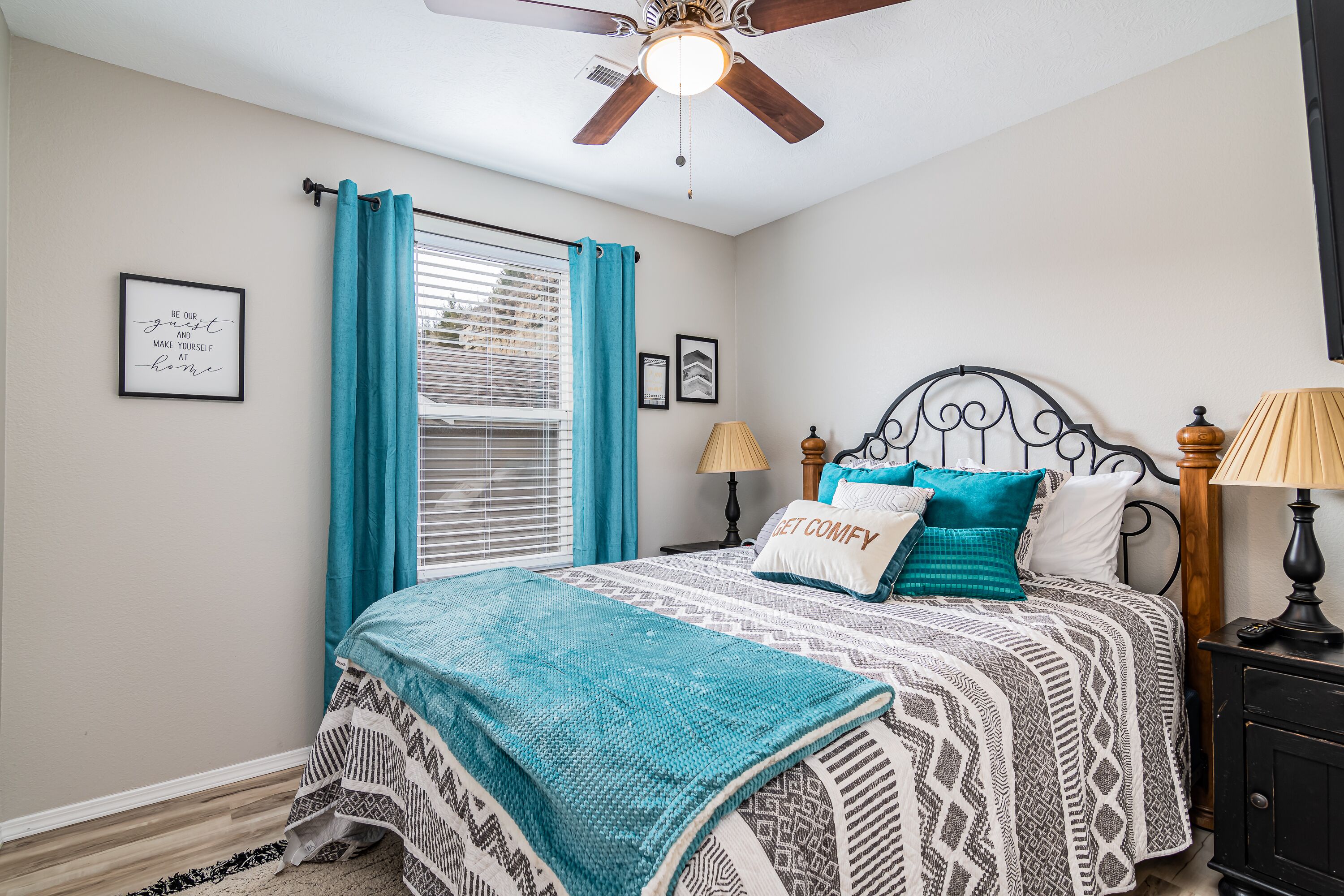 A bedroom with a bed, a ceiling fan, a window with curtains, a bedside table with a lamp, and a wall with framed pictures.