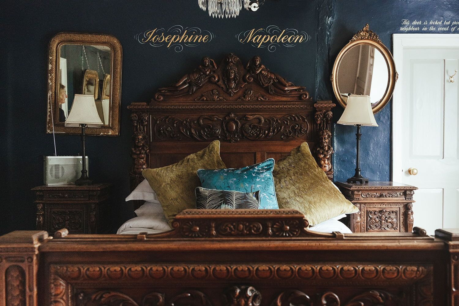 A bedroom with a dark blue wall, a wooden headboard, and a bed with multiple pillows. There are two bedside tables with lamps and a mirror. The room has a chandelier on the ceiling.