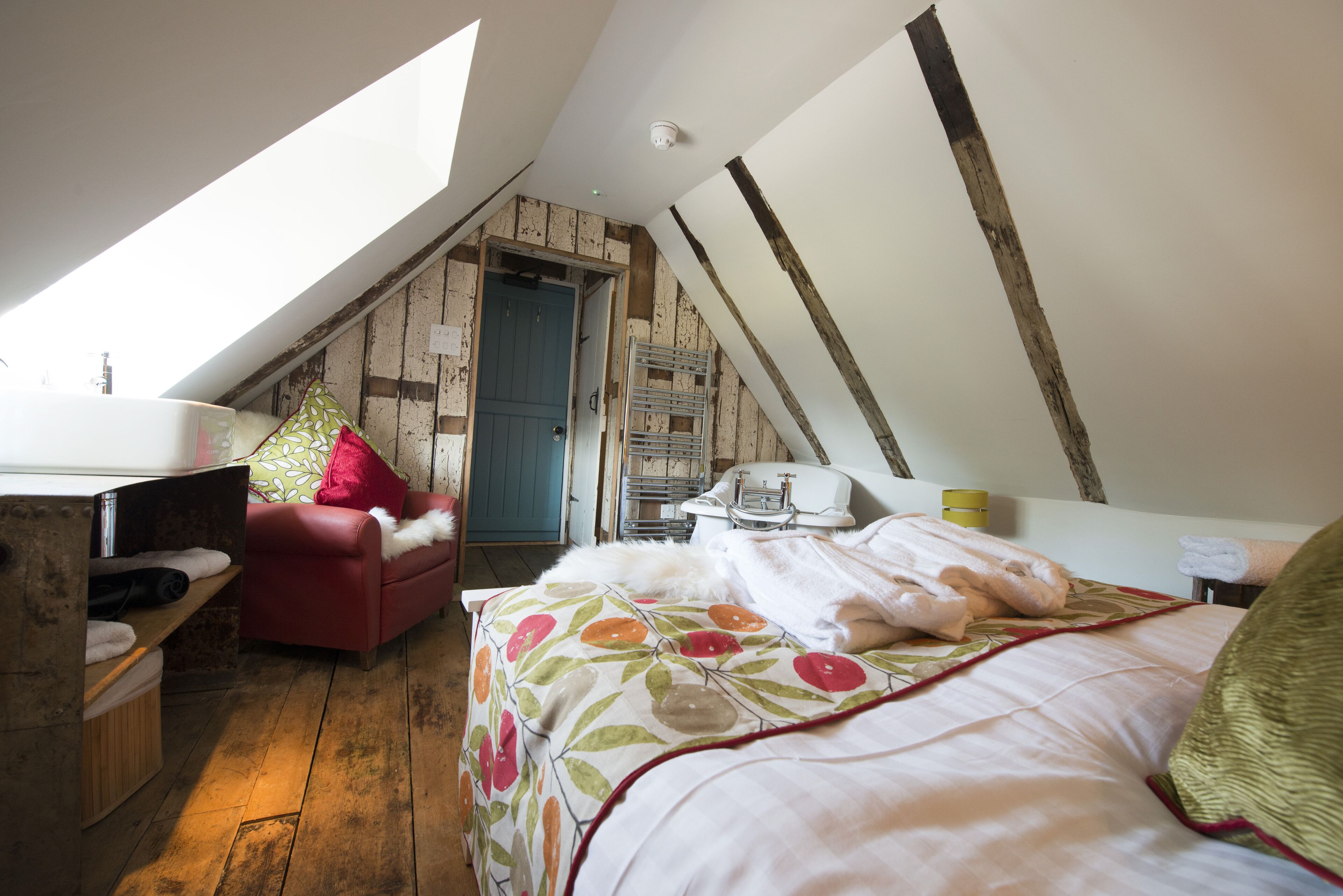 Ein gemütliches Schlafzimmer im Dachgeschoss mit schrägen Wänden, einem roten Sofa, einem Bett mit floralem Bettzeug und Holzboden.
