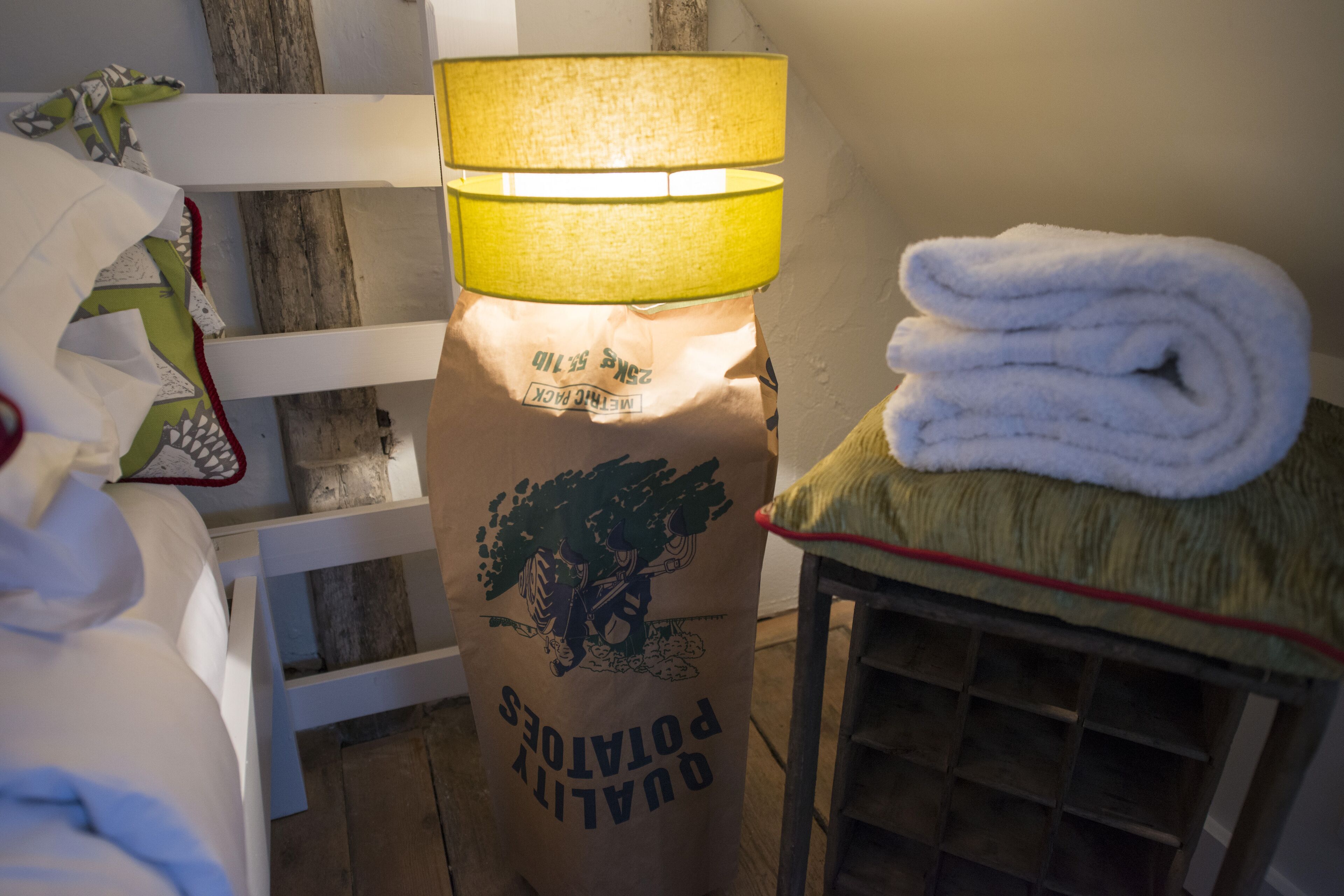 A room with a bed, a lamp, a folded towel, and a bag labeled "QUALITY PORTIONS".