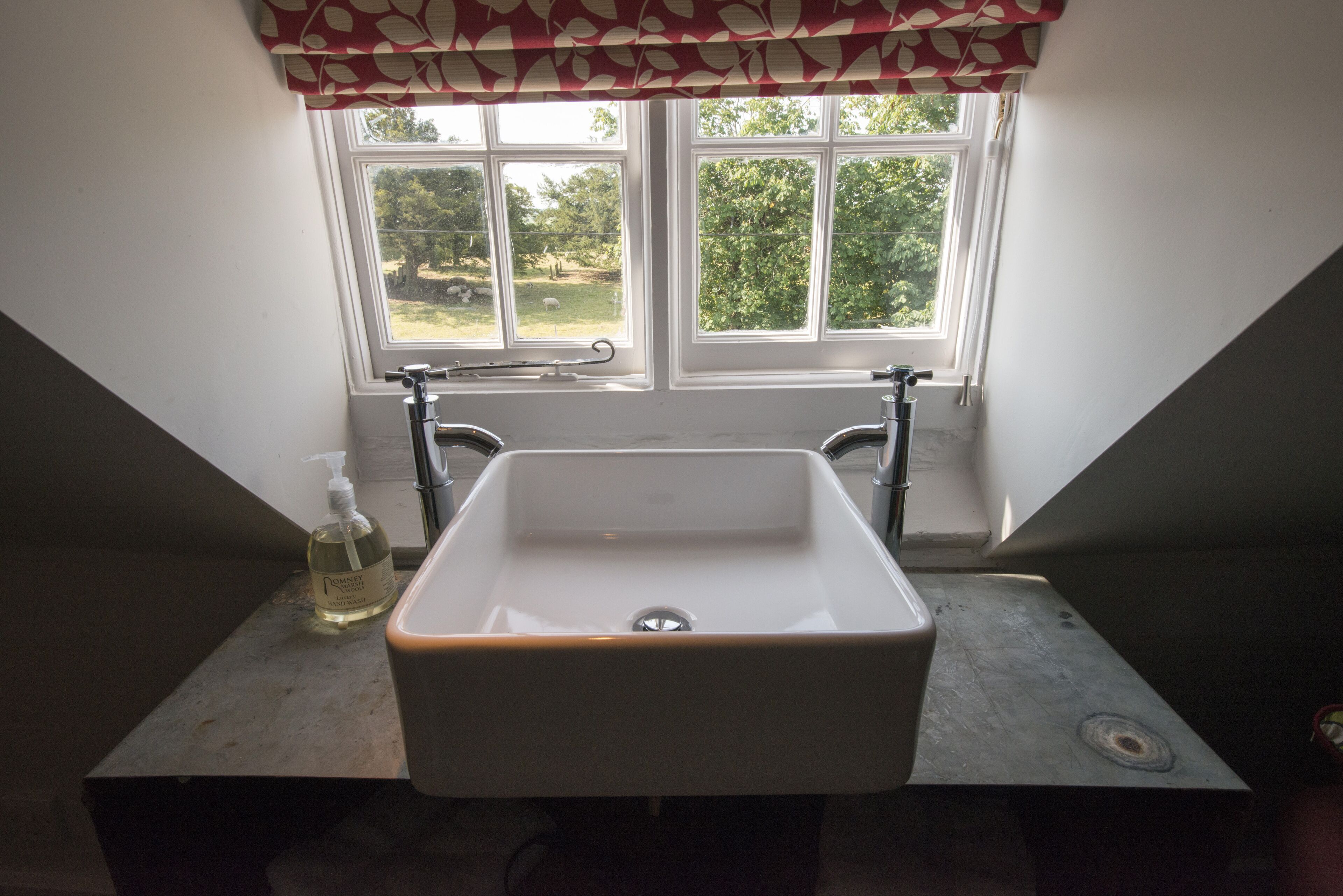 Un baño con un lavabo blanco, una ventana con cortinilla roja y blanca, y vistas al exterior con vegetación.
