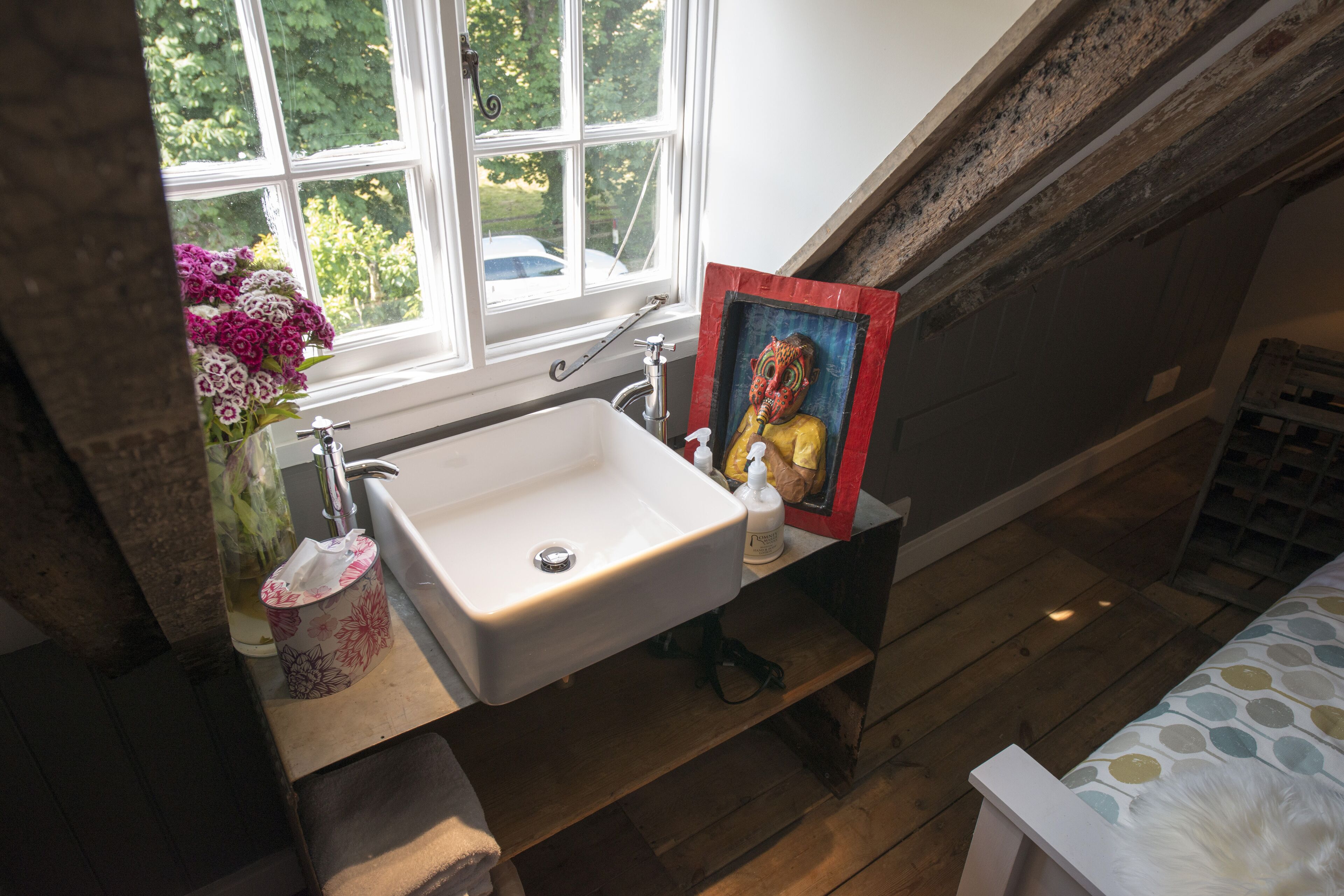 A bathroom with a white sink, a window with a view of greenery, and a painting on the wall.