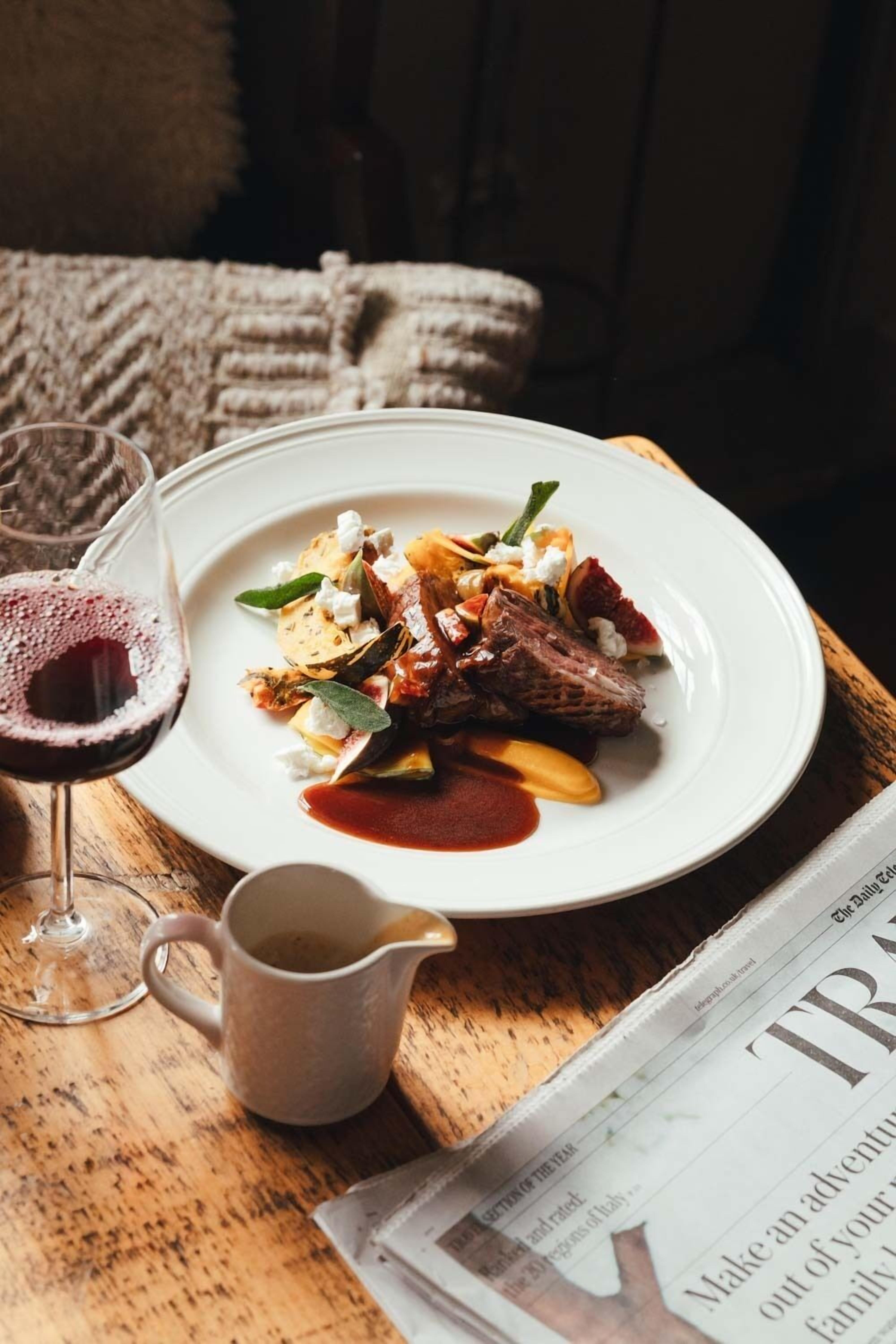 A plate of food with a glass of red wine and a newspaper on a wooden table.