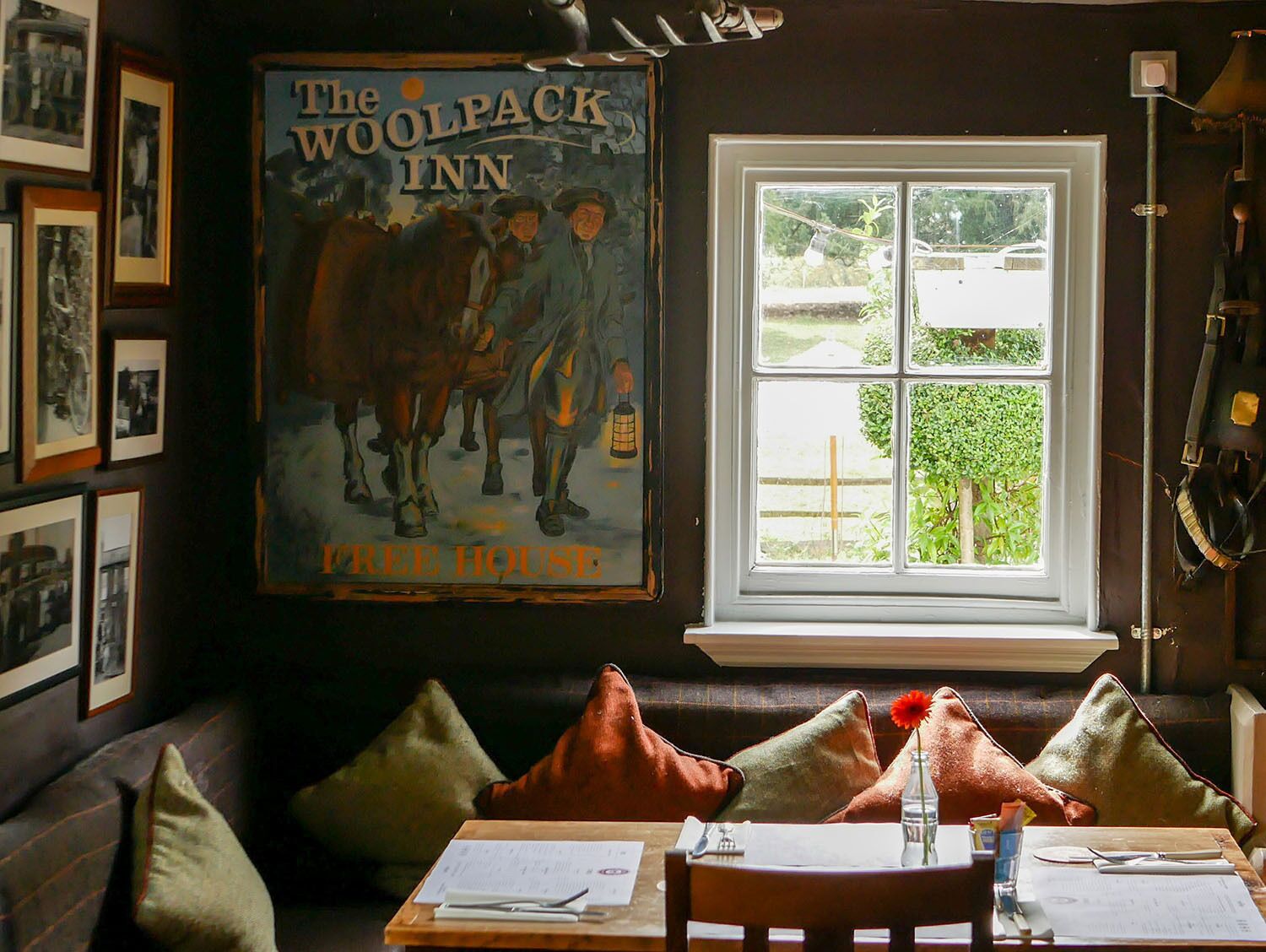Una habitación acogedora con un cartel grande de 'The Woolpack Inn' y 'Tree House' en la pared, una ventana con vistas a vegetación, y una mesa con una botella y papeles.
