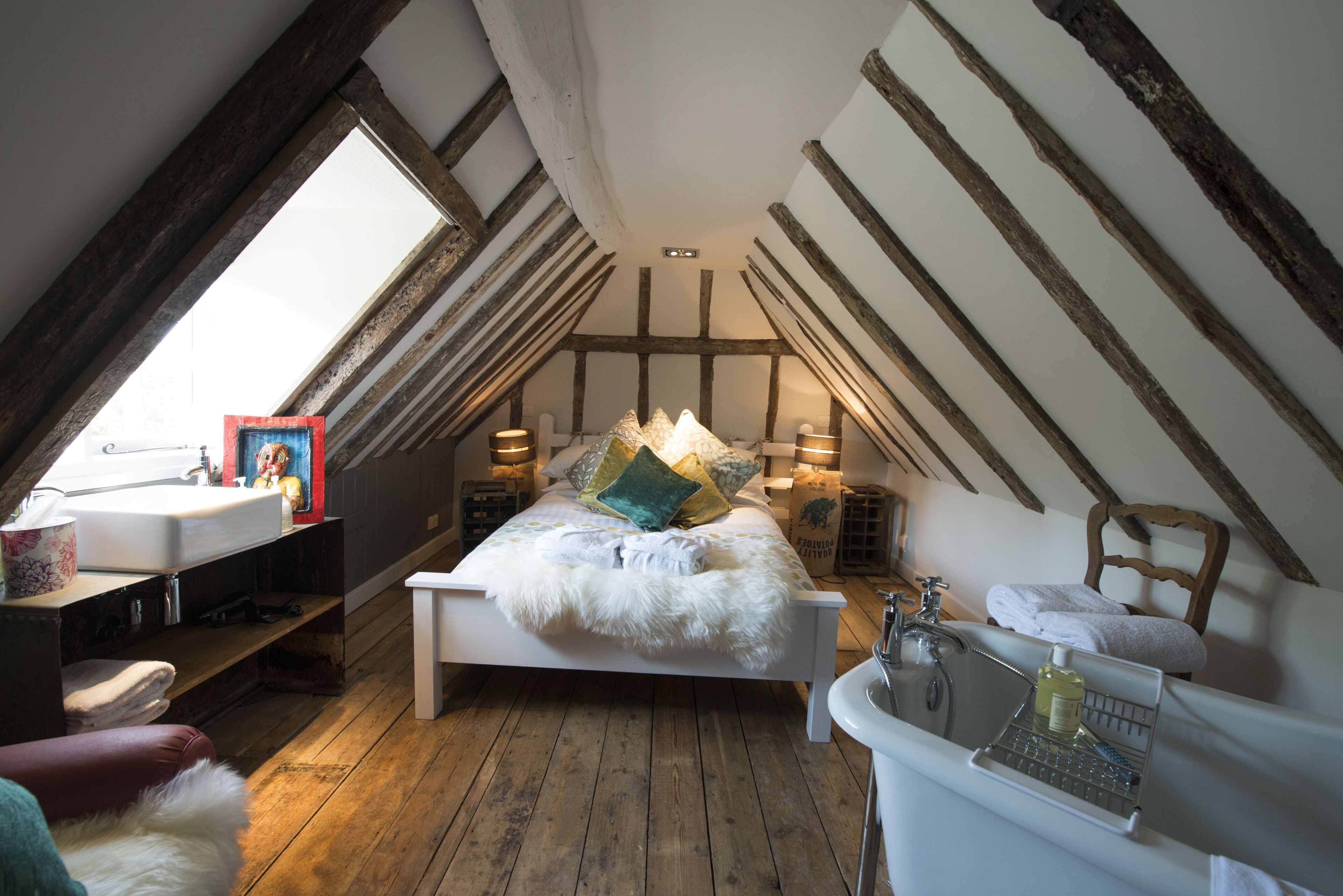 Un dormitorio con techo inclinado, una cama con almohadas, una mesita de noche con lámpara, una ventana y un baño con lavabo y espejo.