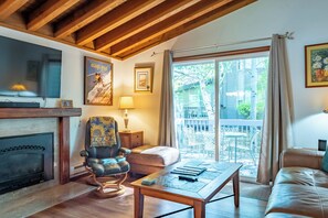 A cozy living room with a fireplace, a comfortable armchair, a coffee table, and a large window offering a view of the outdoors.