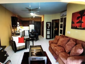 A living room with a brown sofa, a coffee table, and a ceiling fan. There is a kitchen area in the background with cabinets and a microwave.