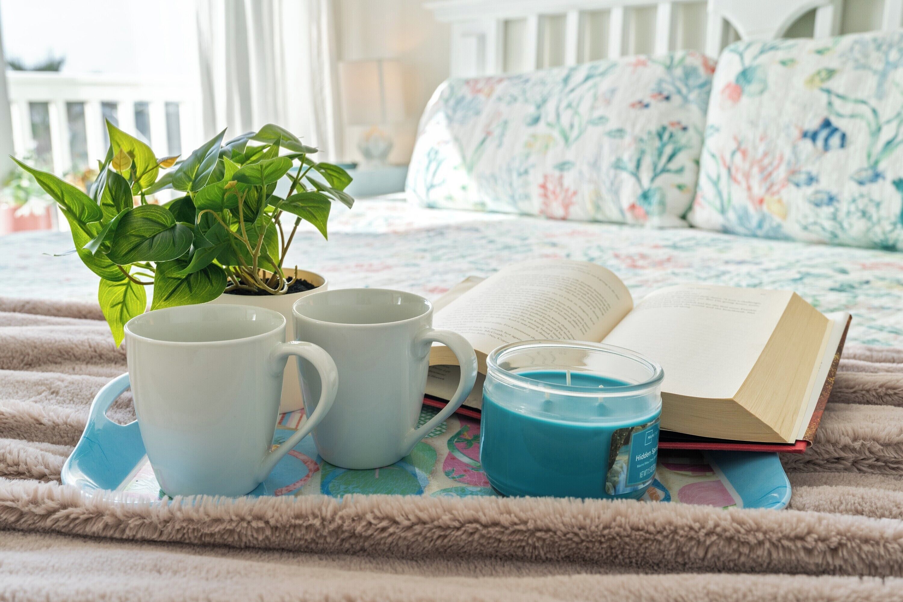 A cozy bedroom with a bed, a potted plant, two white mugs, a blue candle, and an open book on a fluffy blanket.