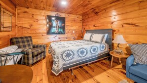 Habitació d'una cabana de fusta amb un llit, un butaquesa escocès, un sofà blau, una taula petita amb una làmpada i una televisió penjada a la paret.