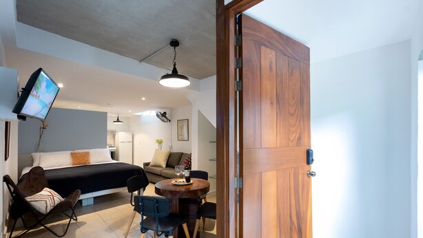 A modern room with a bed, a dining table with chairs, a TV mounted on the wall, and a wooden door.