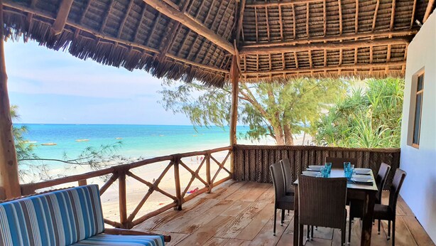A wooden deck with a thatched roof overlooking a beach. There are striped chairs and a table with blue glassware.