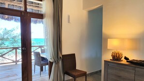 A room with a wooden floor, a chair, a desk with a lamp, and a view of the sea through a glass door.