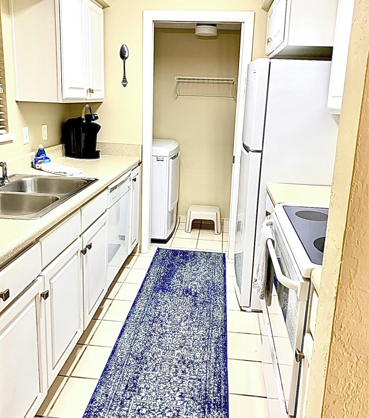Une cuisine de taille réduite avec des meubles blancs, un évier en acier inoxydable, un réfrigérateur blanc et un tapis à motifs bleus.