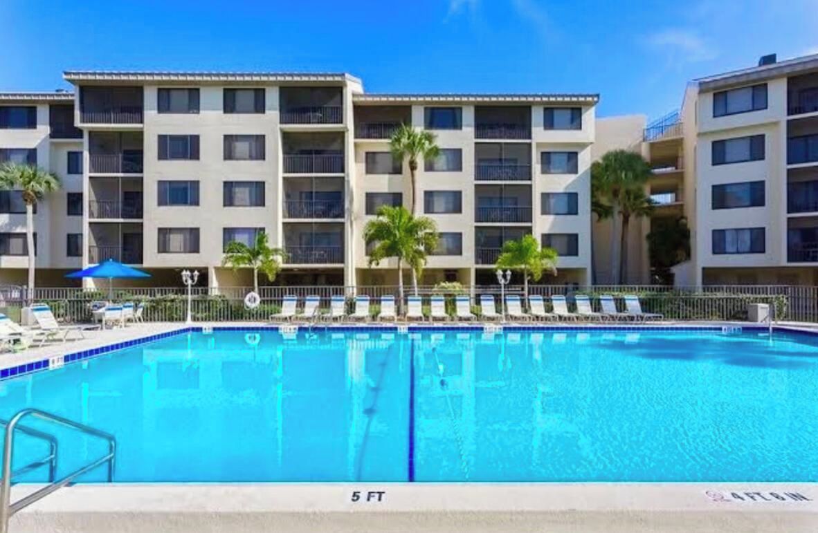 A swimming pool with a 5 ft depth, surrounded by a building with multiple balconies and palm trees.