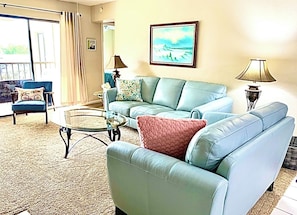 A living room with a sofa, armchair, and coffee table. There is a painting on the wall and a lamp on a side table.