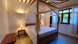 Een kamer met een hemelbed, een houten bankje, een nachtkastje met een lamp en een raam met gordijnen.