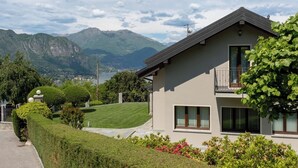 A modern two-story house with a balcony, surrounded by well-maintained gardens and a mountainous backdrop.
