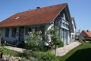 A two-story house with a red-tiled roof, a balcony on the second floor, and a garden with various plants and flowers.