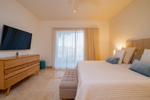 Quarto com cama, penteadeira de madeira, televisão e janela com cortinas.