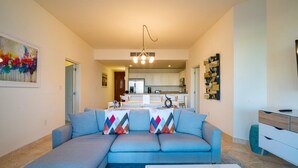 A modern living room with a sectional sofa, a coffee table, and a kitchen in the background. - Paraiso Del Mar Resort E202 2 Bed by Casago (La Paz)