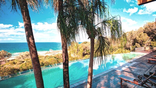 Zona junto a la piscina con vista al océano, rodeada de palmeras y una terraza de madera.
