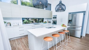 Una cocina moderna con armarios blancos, una isla grande con taburetes, electrodomésticos de acero inoxidable y vistas al verde a través del ventanal.