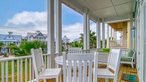 Une véranda couverte, avec des chaises et une table en bois peintes en blanc, donnant sur un quartier résidentiel avec des maisons et des palmiers.