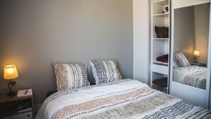 Un dormitorio con una cama, dos almohadas, una mesita de noche, una lámpara y un armario con espejo.