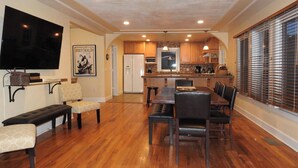 A spacious living area with a wooden floor, a dining table with chairs, a bench, a flat-screen TV mounted on the wall, and a kitchen in the background. - 1927 - Murphys Vacation Rentals (Murphys)