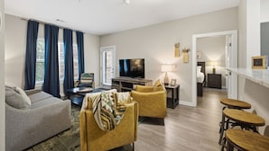 Un salon moderne avec un canapé, un fauteuil et un téléviseur. On y trouve également un coin repas avec une table et des chaises. La pièce est décorée dans des tons neutres et est éclairée par un luminaire au plafond.