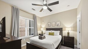Une chambre à coucher comprenant un lit, des tables de chevet, un bureau, un ventilateur de plafond et une fenêtre avec des stores.